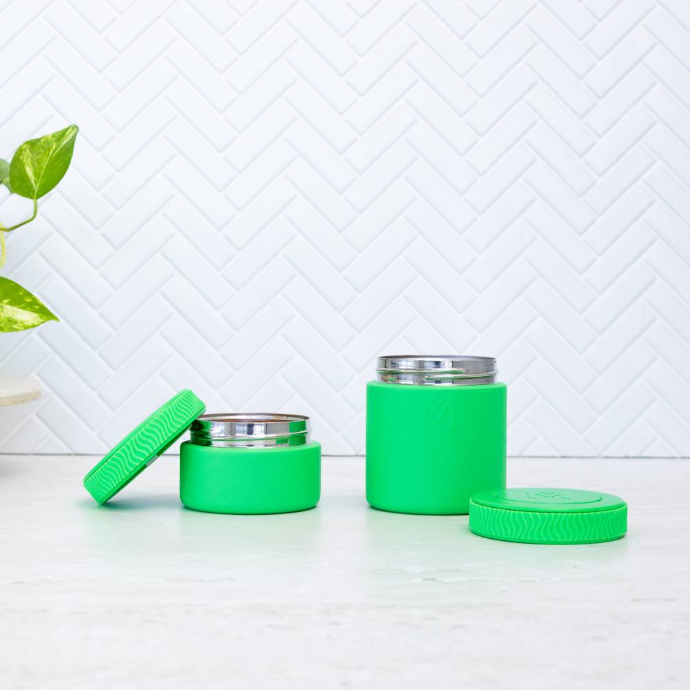 Two green containers with silver lids on a white surface with a white geometric patterned wall in the background.