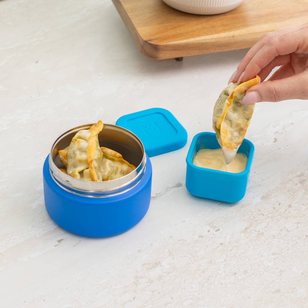 Blue food container with a hand dipping a dumpling into a small bowl of dip on a marble surface.