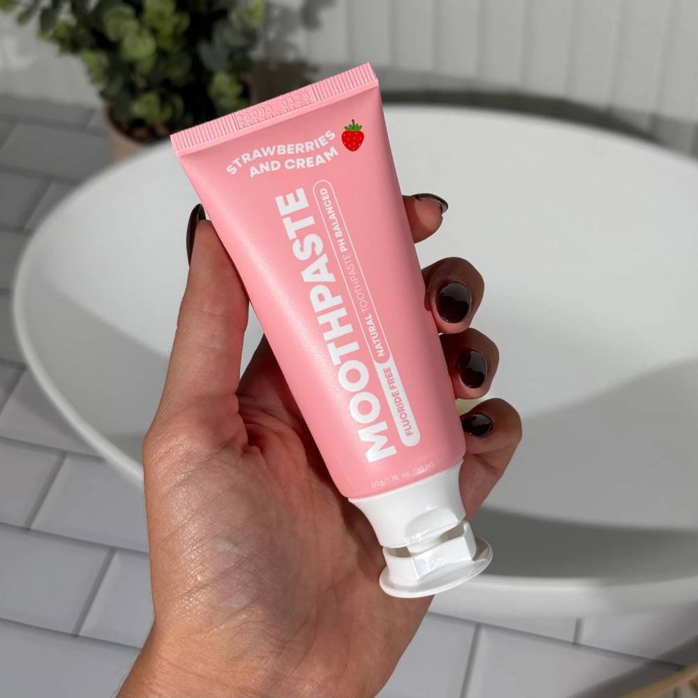 A hand with dark manicured nails holds a pink squeeze tube of Moothaste Strawberry and Cream toothpaste in front of a white sink and tiled bathroom background.