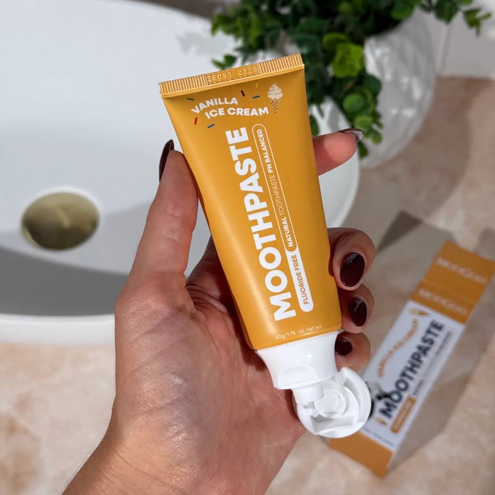 Hand holding a tube of 'MOOTHPASTE' with visible toothpaste, against a bathroom background.