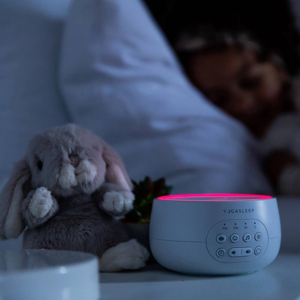 White noise machine with red night light sitting on a bedside table in a dark childrens room.