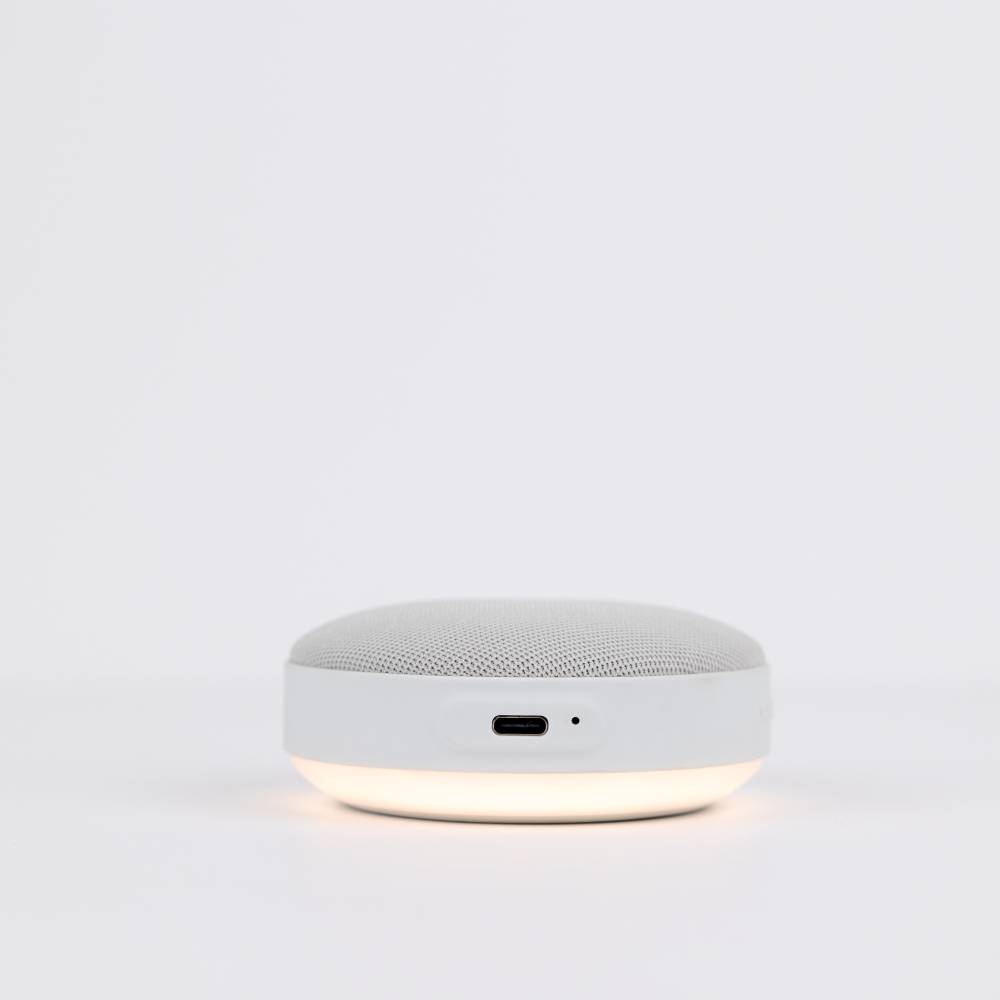 Round white noise machine side view with light on and charging port visible on a light gray background