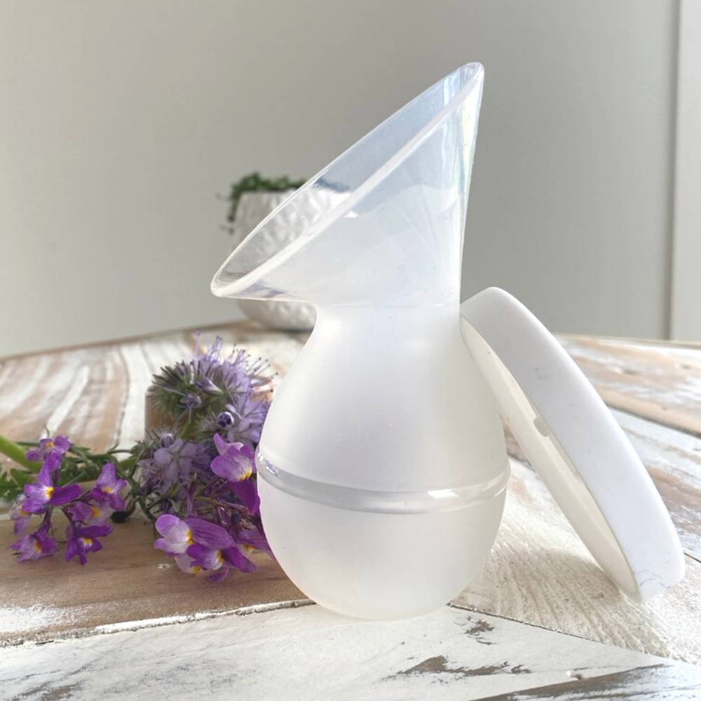 A translucent white silicone breast pump bottle-shaped accessory with a matching white lid, shown on a rustic white wooden surface next to purple flowers.
