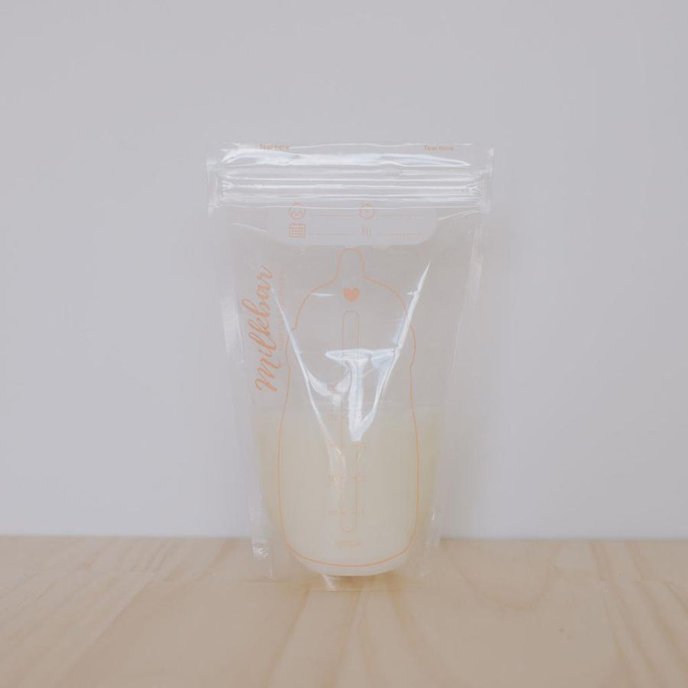 A clear plastic milk storage bag with a resealable zipper, containing pale liquid, standing on a light wood surface against a plain light gray background. The bag has faint peach-colored measurement markings and the word 'Milkbar' printed vertically on the side.