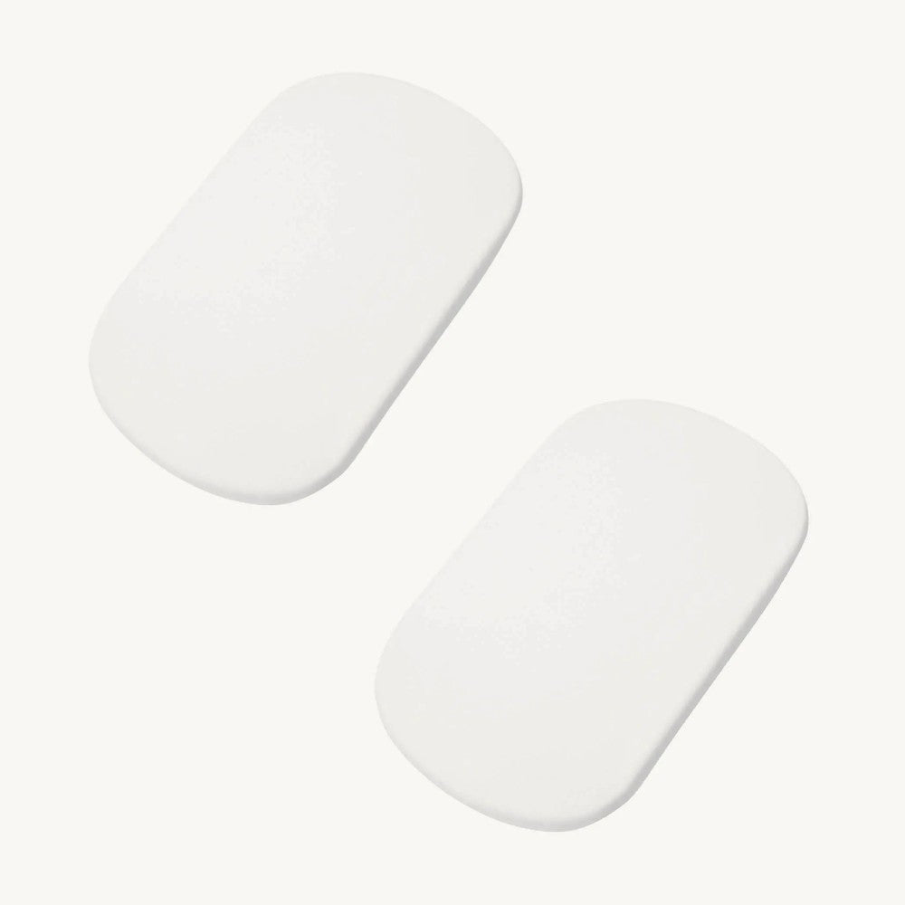 Two white, oval-shaped bassinet mattresses with white sheets, shown on a clean off-white background.