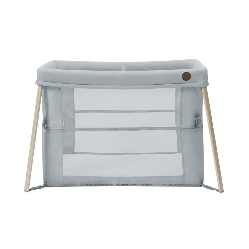 A light gray fabric baby MaxiCosi travel cot with a natural wood frame and clear mesh panels, shown on a solid white background.