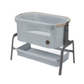 A light gray fabric baby bassinet with a beige trim, mounted on a dark gray metal frame with a lower storage tray, shown against a plain white background.