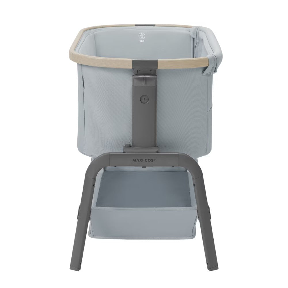 A Maxi-Cosi baby bassinet with a light blue-gray fabric basket, beige trim, and a dark gray metal frame with a matching storage tray below, shown on a solid white background.
