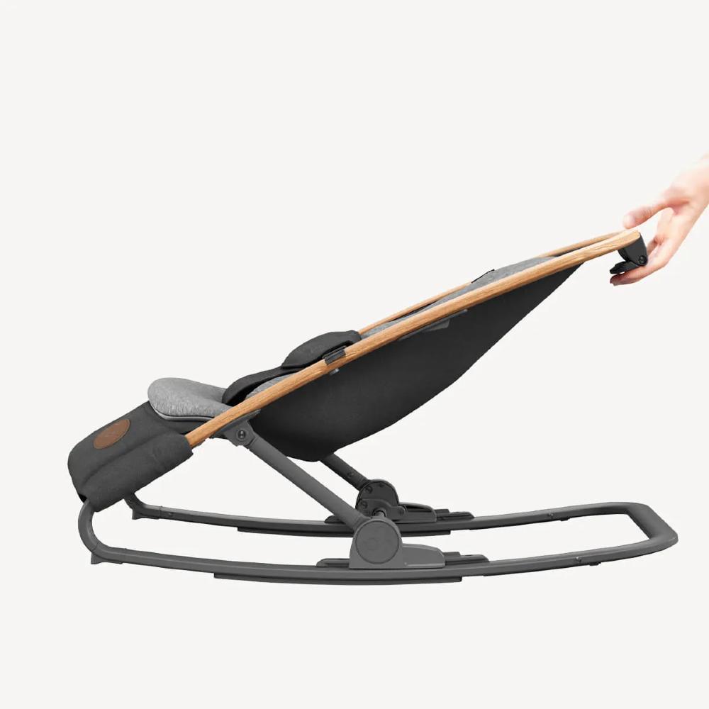 A modern baby bouncer chair with a black fabric seat, light brown wooden handles, and a dark gray metal base, shown with a hand adjusting the handle on a light gray background.