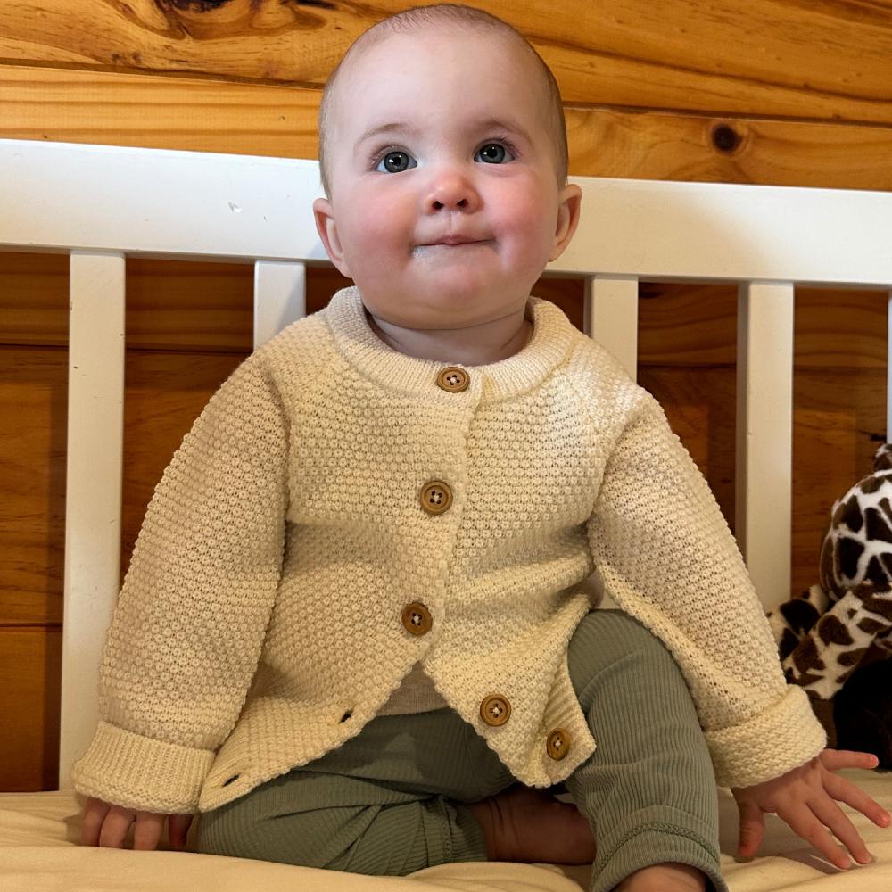 Baby wearing a cream seed stitch wool knitted cardigan with buttons, sitting in a white crib.