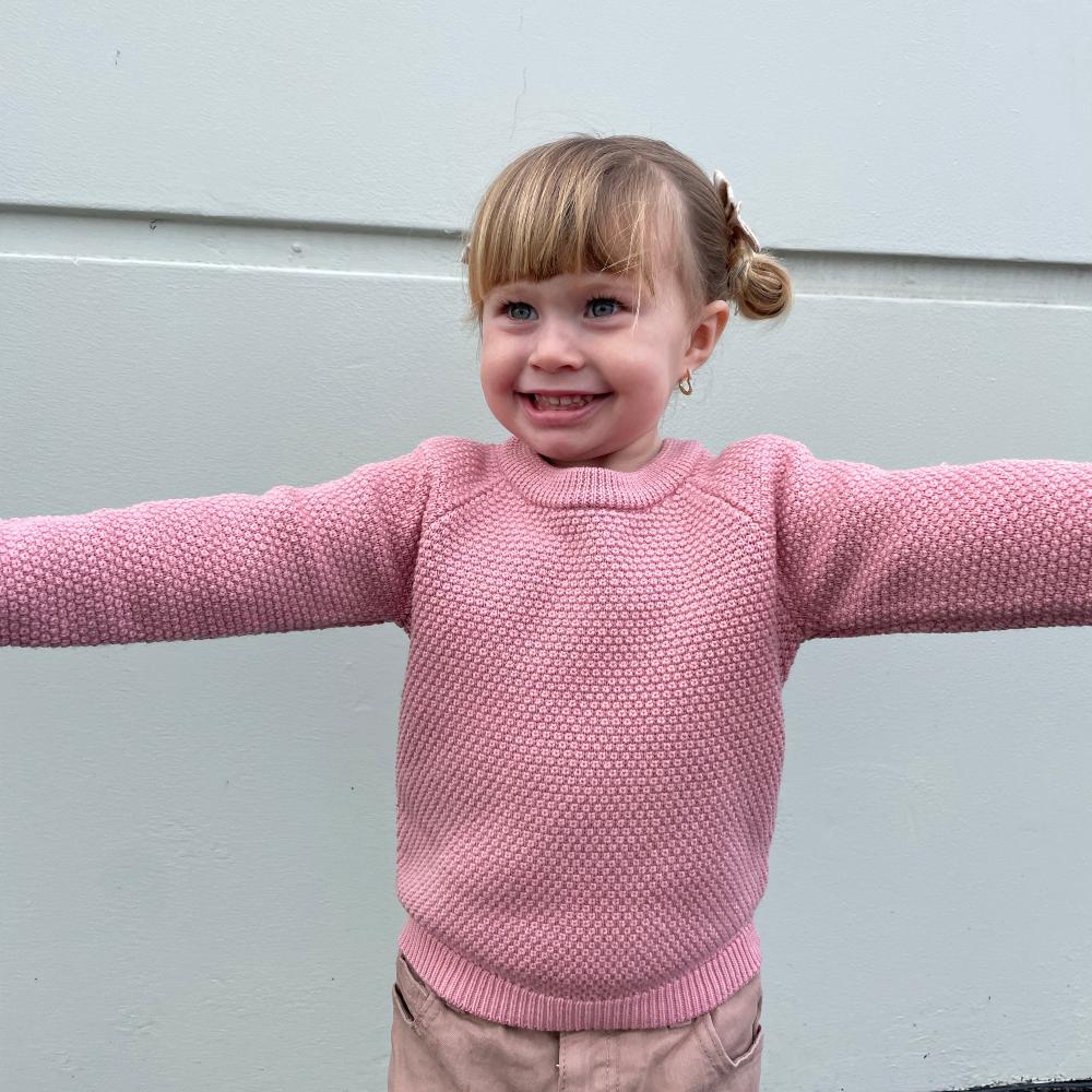 Child wearing a pink seed stitch woolen knitted sweater with arms outstretched against a light gray background