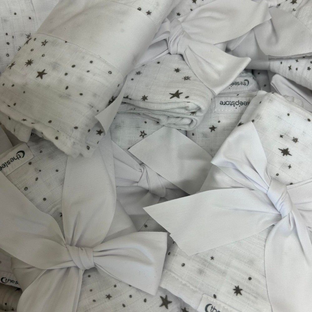 A pile of white muslin baby swaddle blankets decorated with small black star prints and tied with large white satin bows. A brand tag reading 'thesleepstore' is visible on some of the blankets.