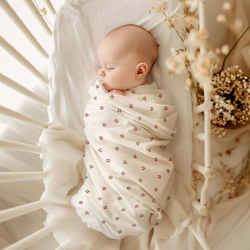 A sleeping baby in a cot wrapped in a stretchy swaddle with a cherry pattern.