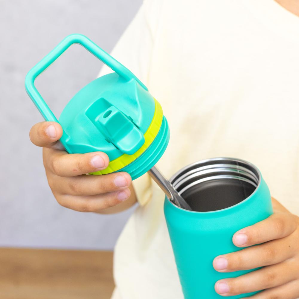 A close-up of a person holding a teal insulated tumbler and lifting its matching lid, which has a yellow accent band and a carrying handle. The person is wearing a cream-colored top, and the background is a soft gray wall with a hint of wooden flooring.