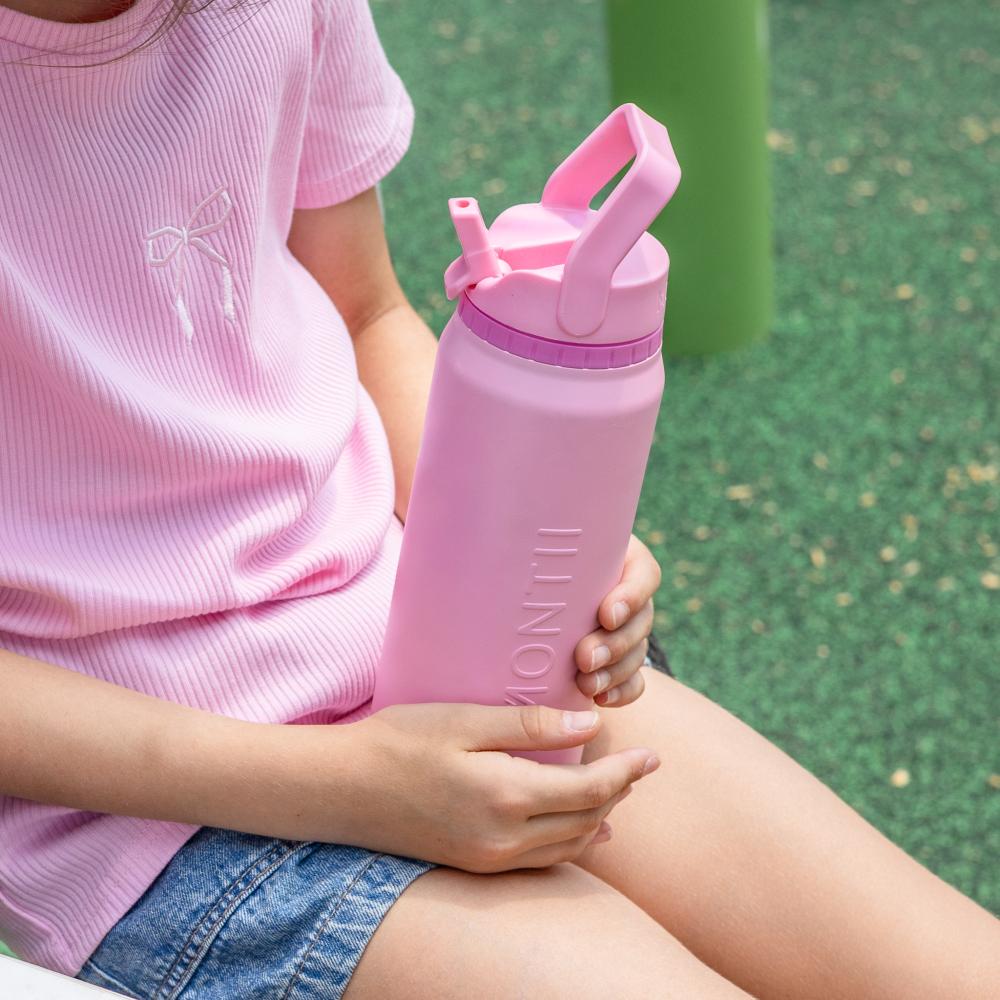 A child in a pink ribbed t-shirt and denim shorts holds a pink MONTI water bottle with a flip-top lid in an outdoor playground setting.