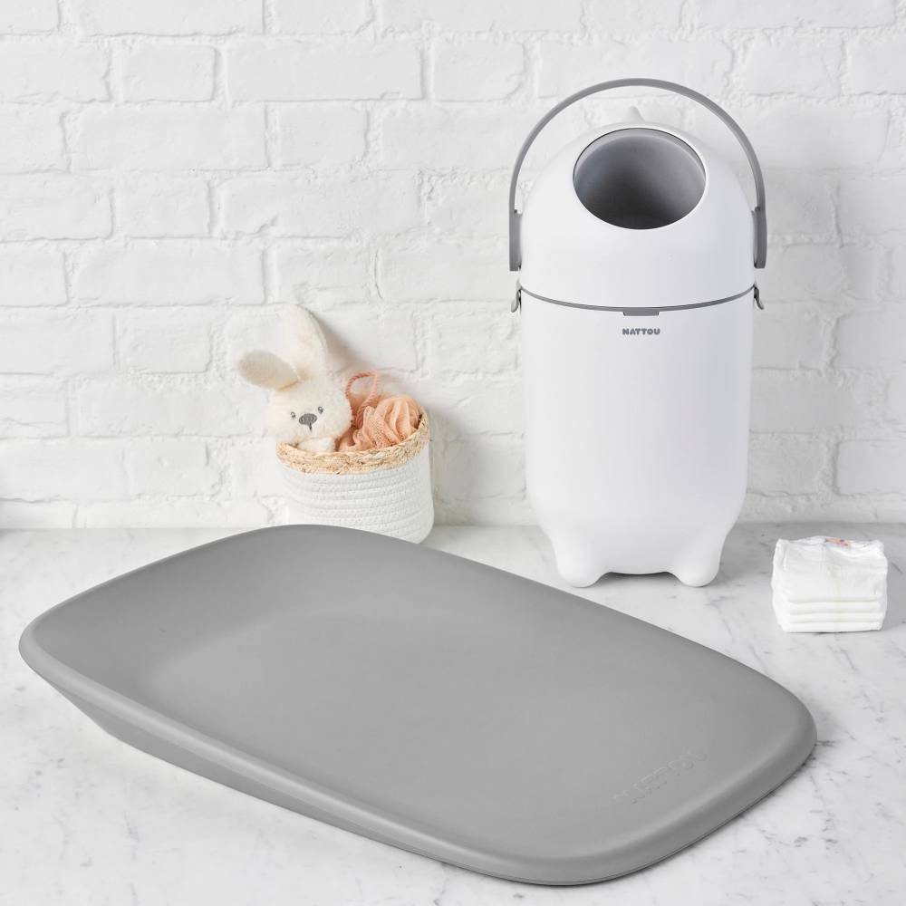 White nappy bin next to grey change table on a light gray surface with a white brick wall background