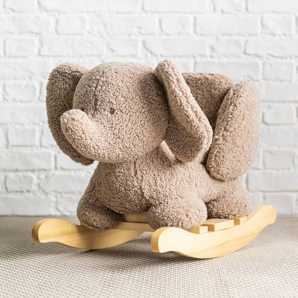 Plush elephant rocking toy on a wooden base against a white brick wall.