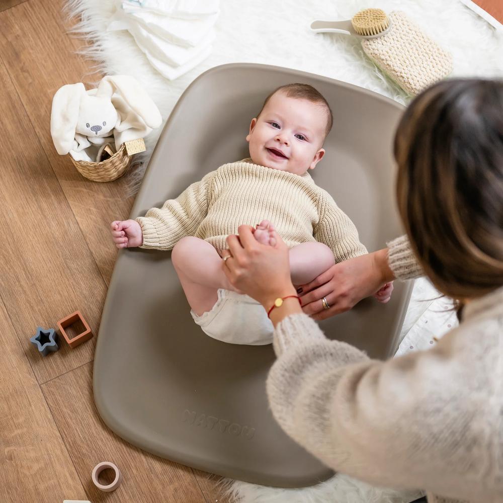 Baby lying on a taupe brown baby change mat with a smiling baby and adult hands, surrounded by toys on a wooden floor.