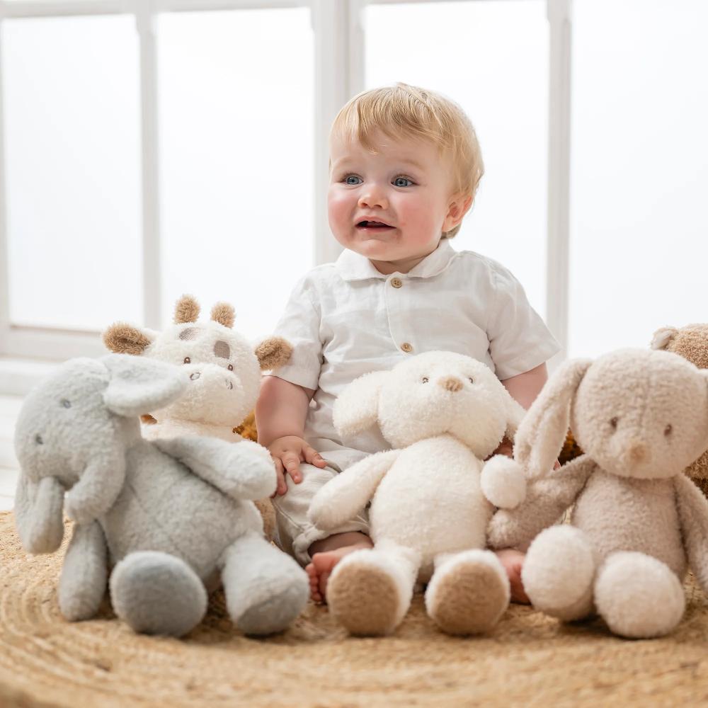 Baby sitting on a carpeted floor surrounded by plush toys in a bright room.