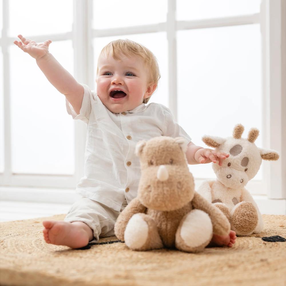 Baby sitting on a carpet with two plush toys, one resembling a bear and the other a giraffe, in a bright room.