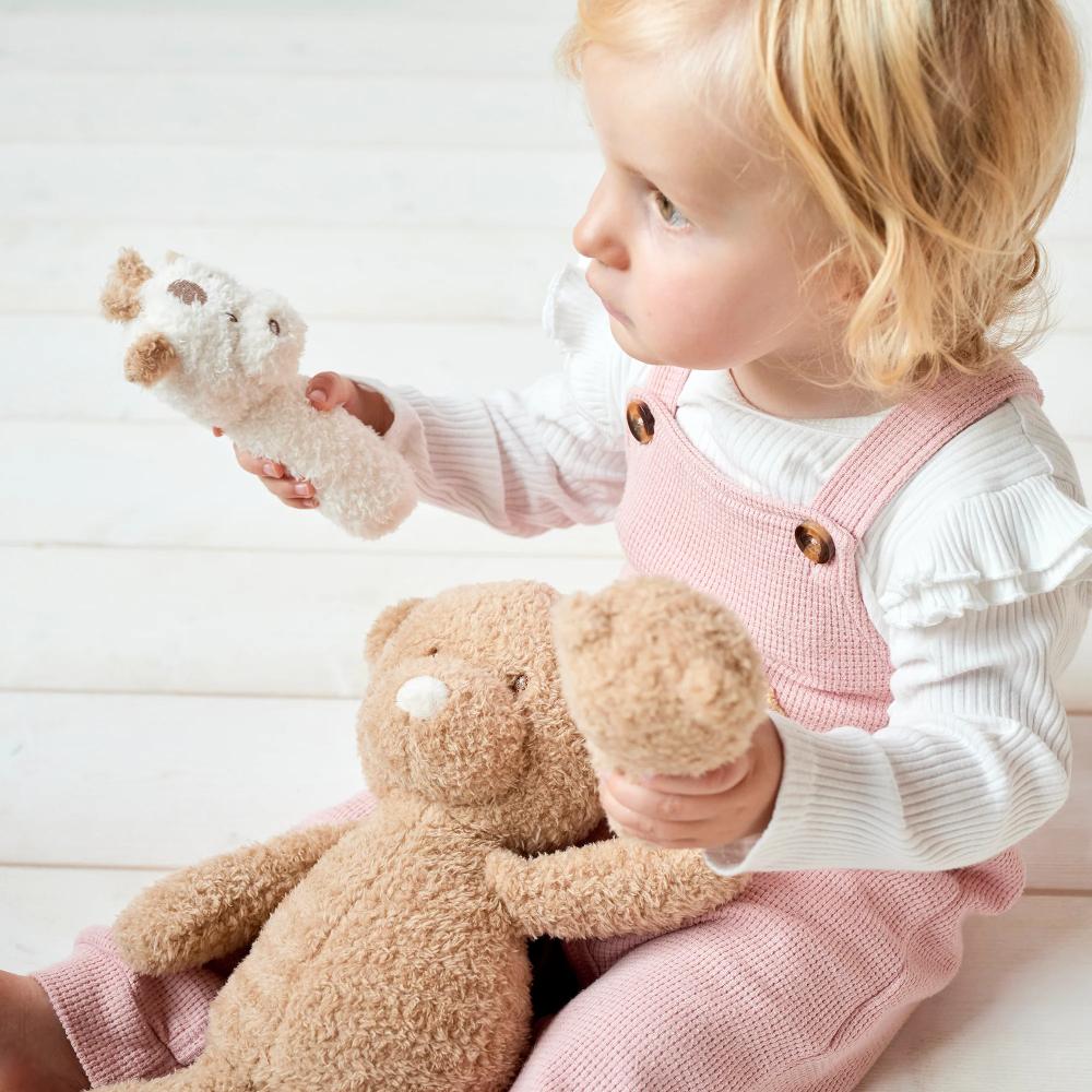 Child in pink overalls holding a plush Nattou rattle on a light wooden floor.