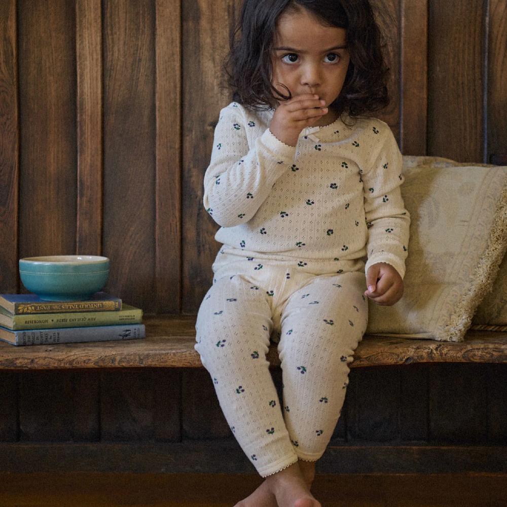 Child in cream and berry print pyjamas sitting on a wooden bench with a bowl and books in the background