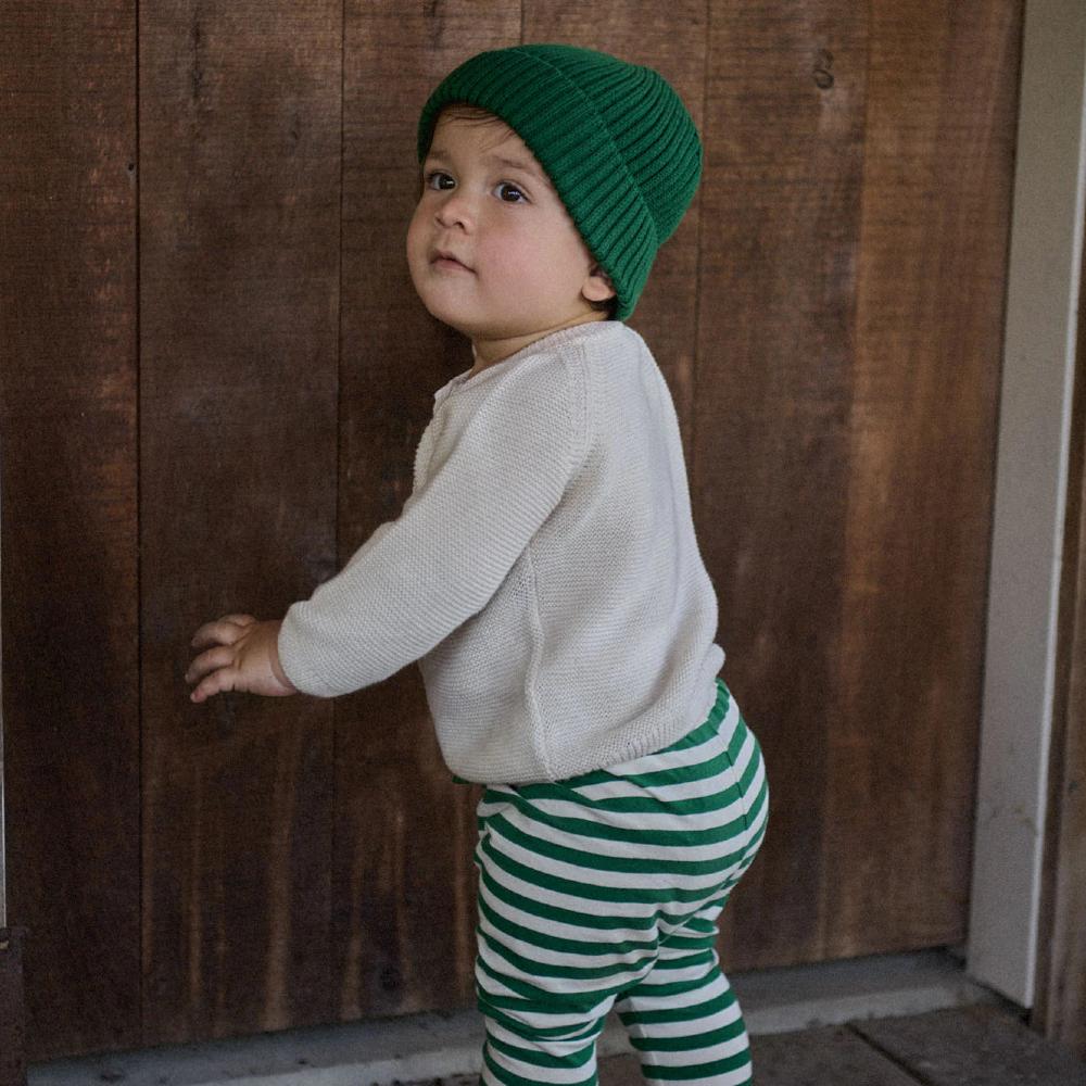 Child wearing a green knit hat, beige sweater, and green and white striped pants standing against a wooden wall.