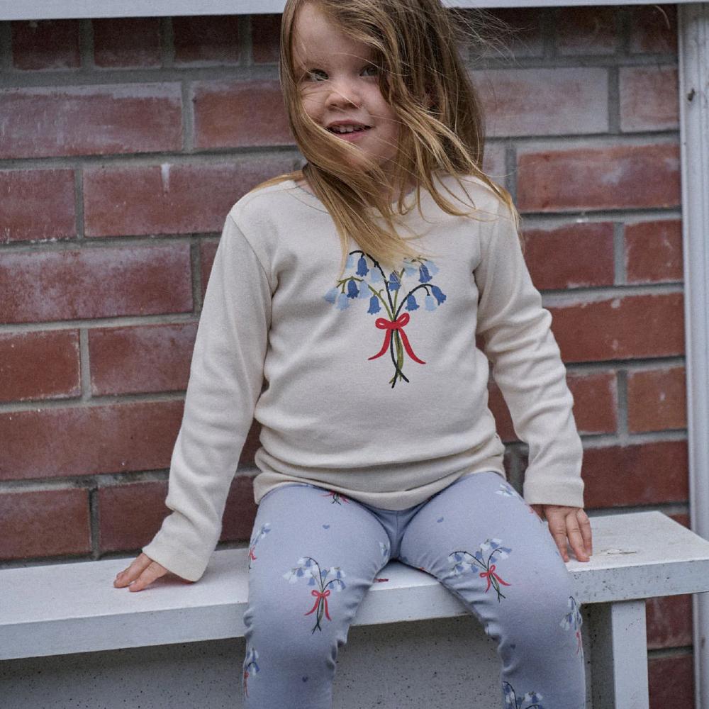 Child wearing a light-colored sweater with a floral design and matching blue pants, sitting on a bench against a brick wall.