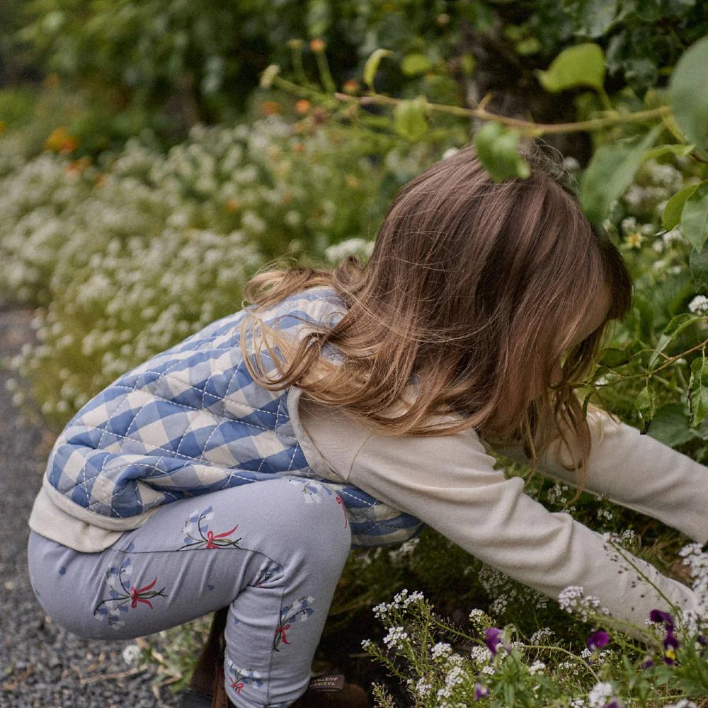 A child playing in a garden with flowers and greenery wearing a blue check vest and floral blue pants 
