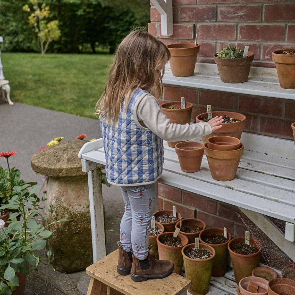 Child arranging potted plants on a table outdoors wearing a check blue vest and blue floral leggings with brown boots