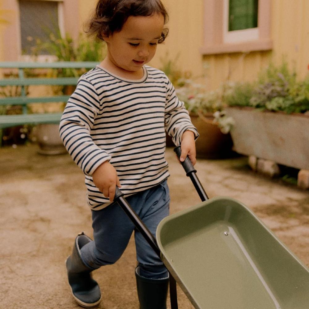 Child in a navy/white striped shirt and rain boots pushing a green wheelbarrow outdoors.
