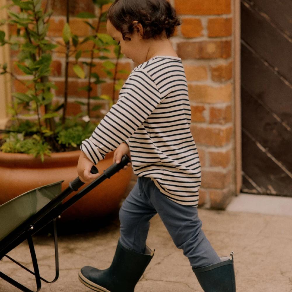 Navy and white child in striped shirt and rain boots pushing a green wheelbarrow outdoors against a brick wall 