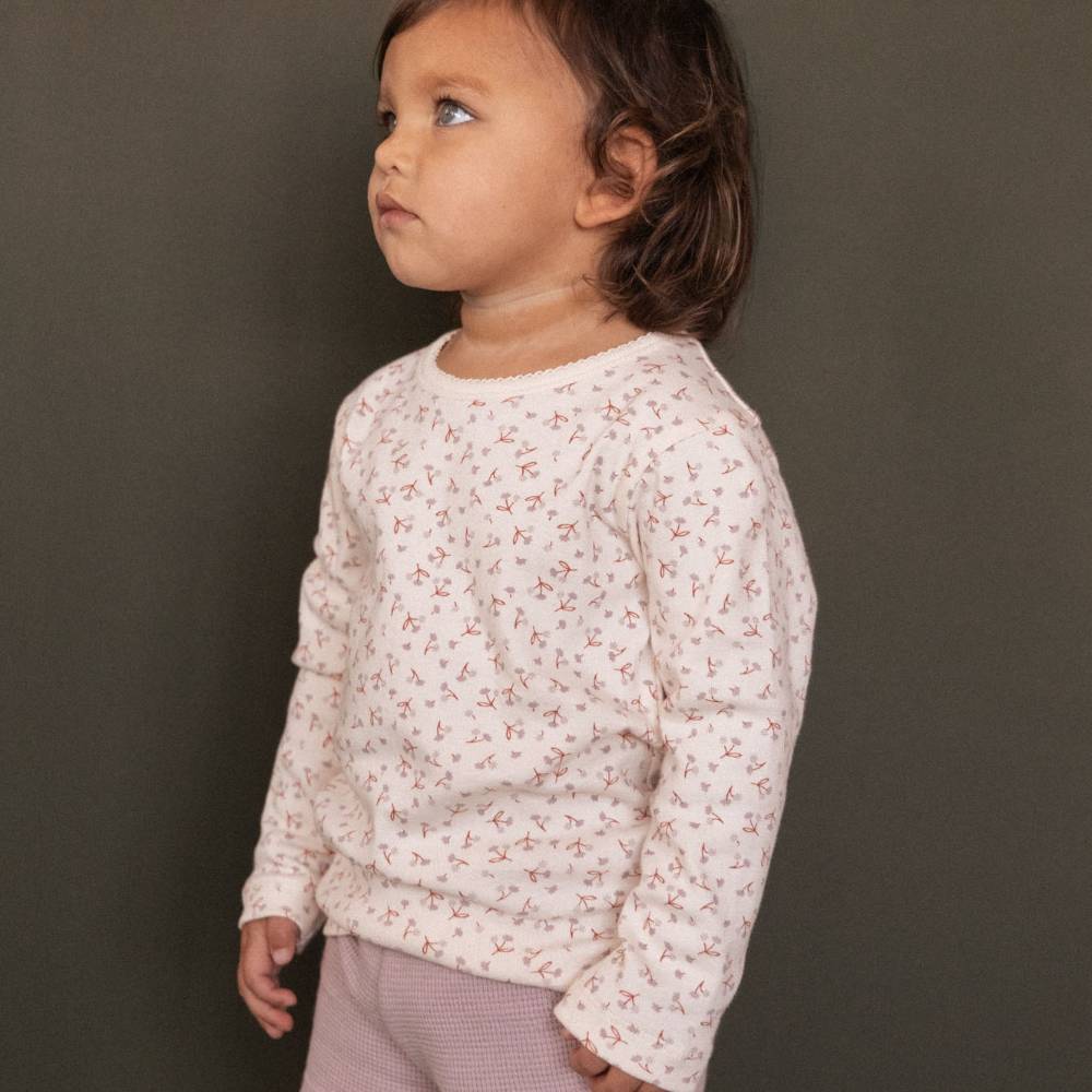 A toddler girl in profile wearing a cotton cream-colored long-sleeve crewneck top with a tiny pink and red floral pattern, paired with light pink pants, set against a solid dark gray background.