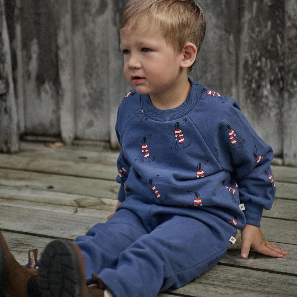 Child wearing a blue sweater with red and white dog patterns and blue trackpants sitting on wooden steps.