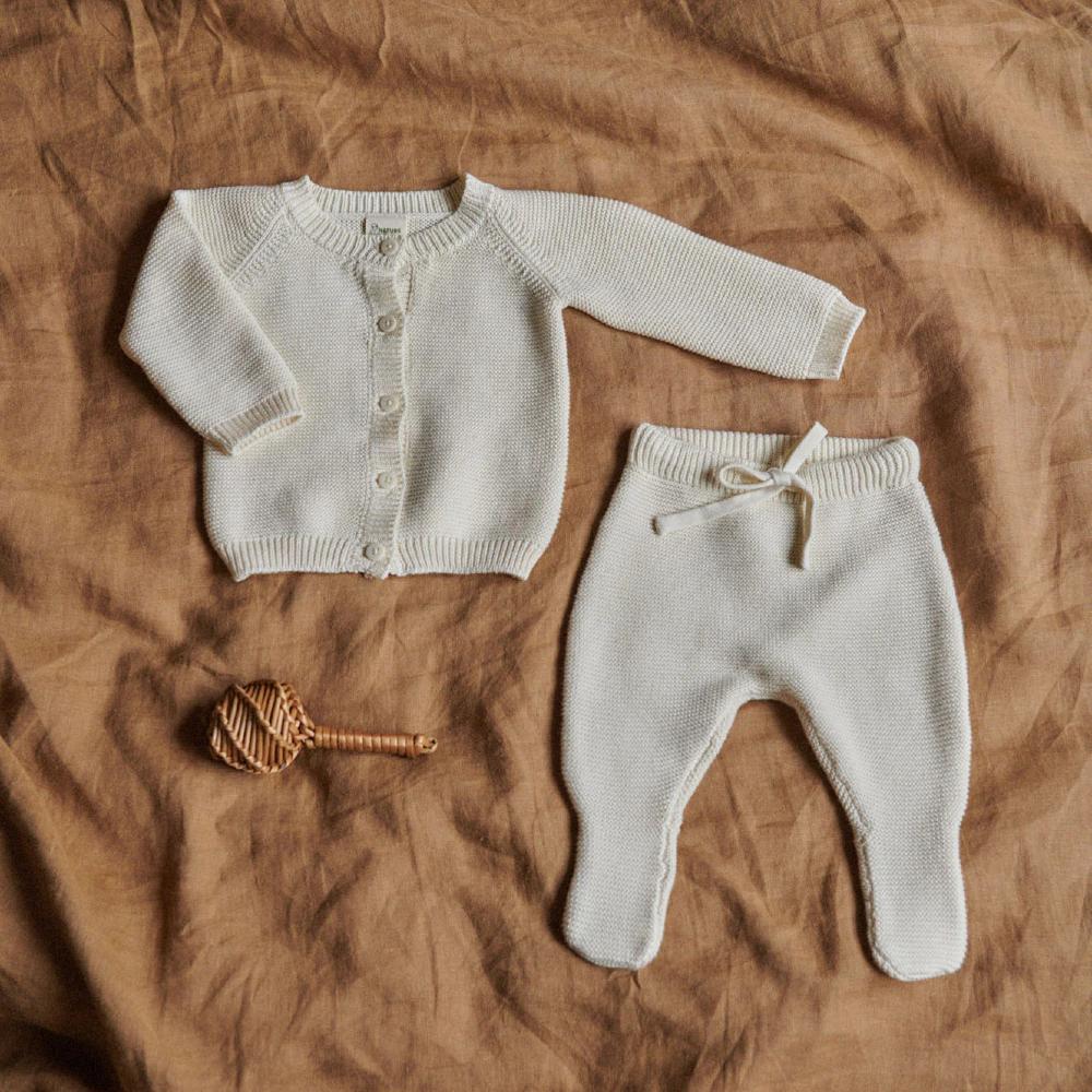 Cream coloured knitted baby cardigan and footed pants with a wooden toy on a brown fabric background