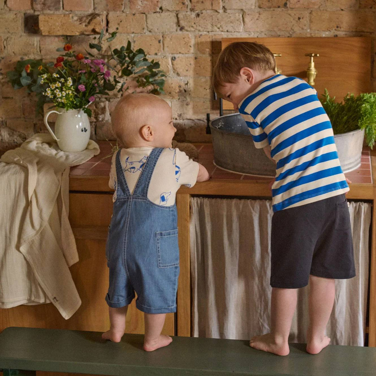 Two toddlers standing on a bench in a rustic kitchen setting, wearing baby clothing. The toddler on the left wears blue denim overalls over a cream shirt, and the toddler on the right wears a blue and white striped shirt with dark shorts.