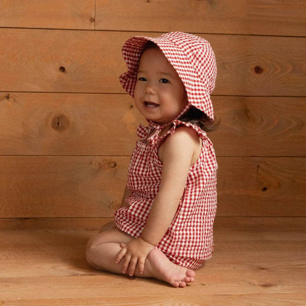 A baby wearing a red and white gingham sleeveless set and matching sun hat, sitting on a wooden floor against a wooden plank wall.