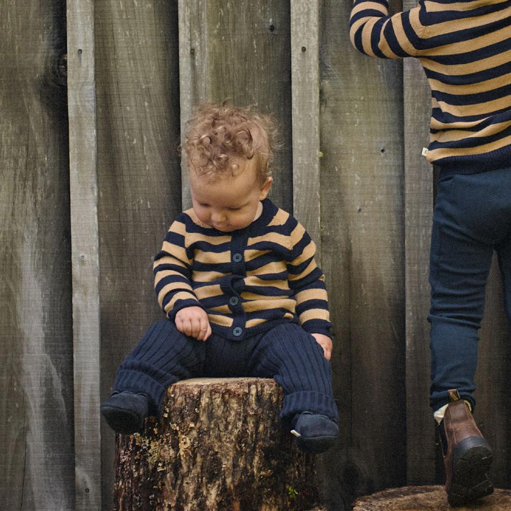 two children in brown and navy striped cardigans and navy blue knitted pants sitting on a log against a wooden fence.