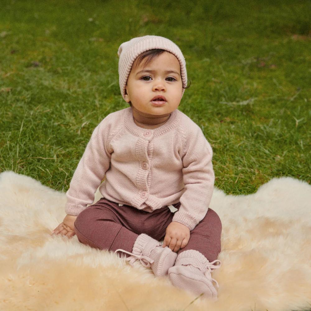 Baby sitting on a fluffy woolen blanket wearing a pink knitted cardigan with a matching hat  and mauve coloured legging with matching pink booties against a grassy background
