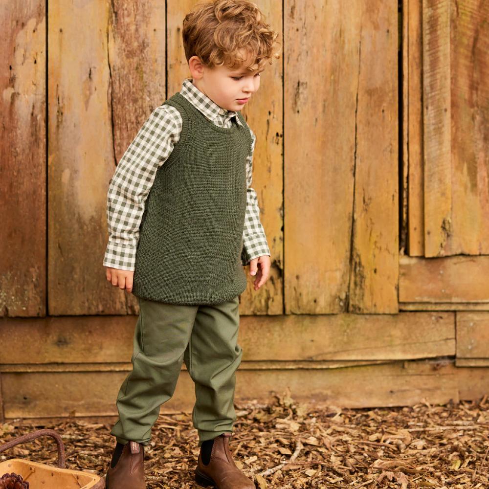 Young boy in a merino wool green knitted vest and plaid shirt standing in front of a wooden wall.