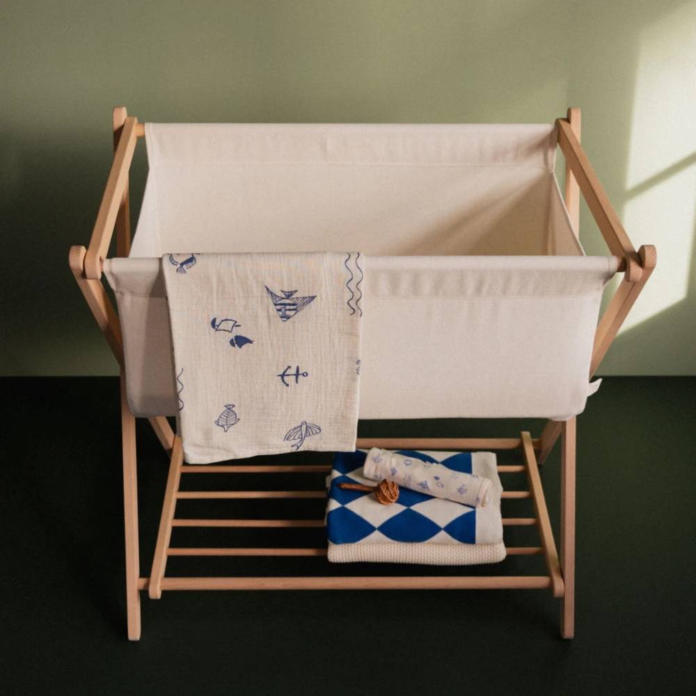 Wooden baby bassinet with cream coloured fabric piece and bright blue and white baby blankets and swaddles arranged on a green background