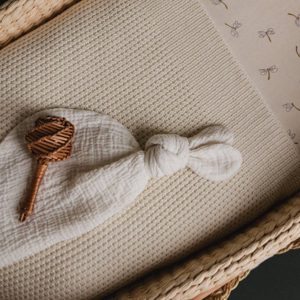 White knotted muslin crinkle texture fabric with a wooden rattle inside a moses basket on a textured knit surface