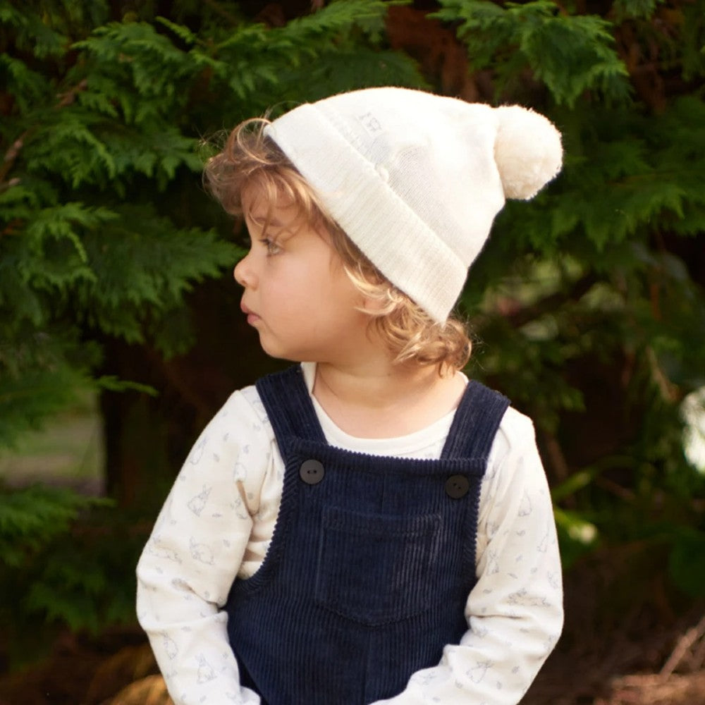 Child wearing a white knit hat with a pom-pom and blue overalls outdoors.