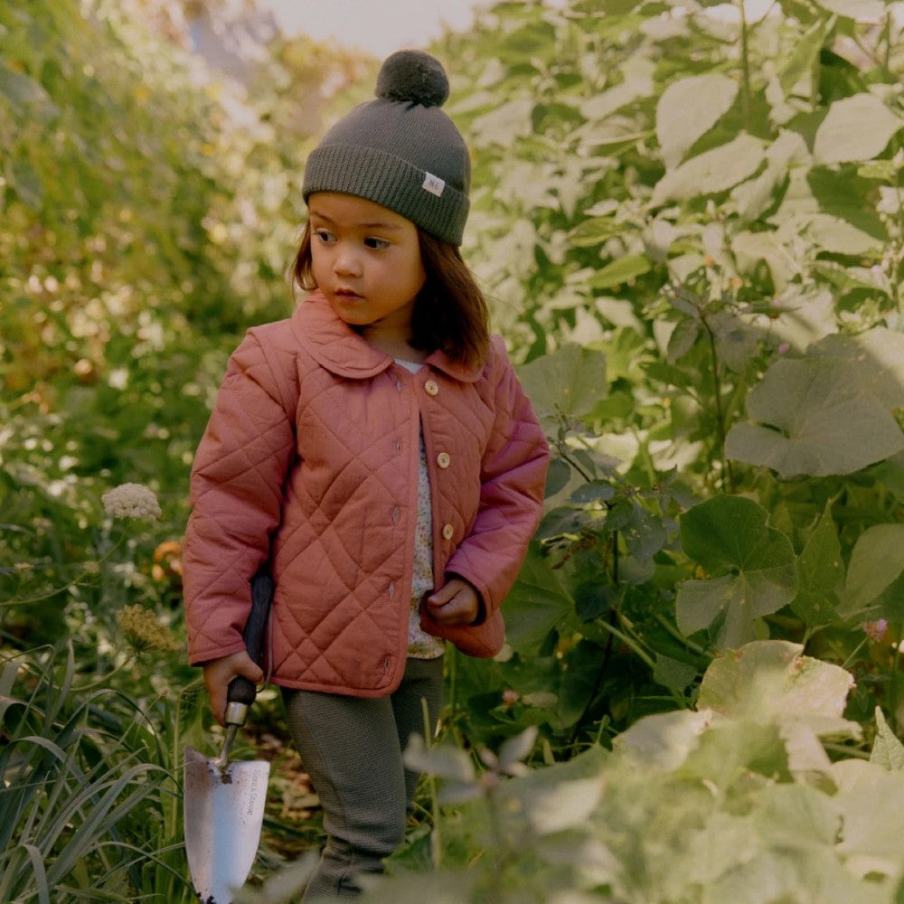 Child in a pink quilted jacket and forest green beanie standing in a garden holding a trowel.