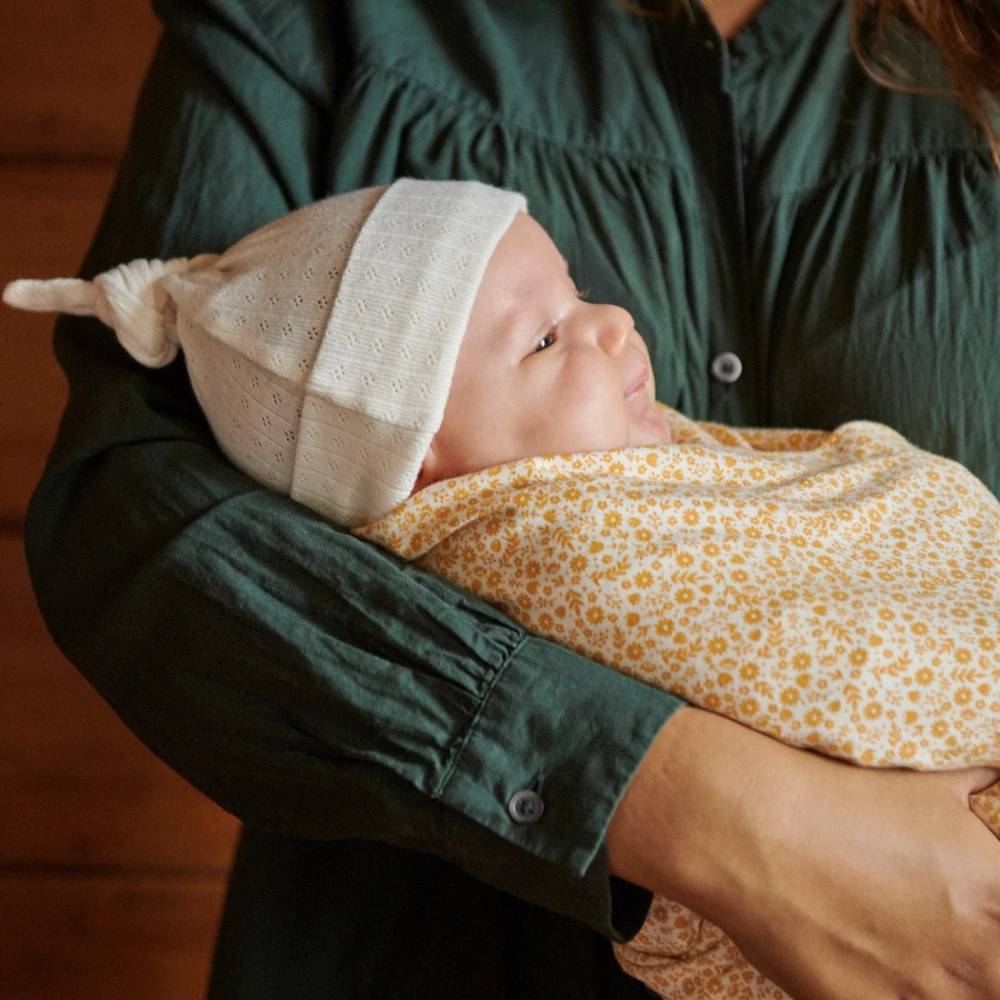 Person holding a baby wrapped in a yellow floral blanket with a white pointelle knit cotton hat with top knot