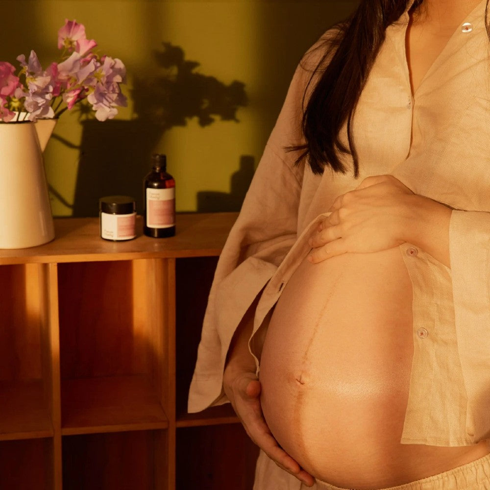 Pregnant person wearing a beige linen shirt with a wooden shelf in the background holding Nature Baby belly oil and cream containers 