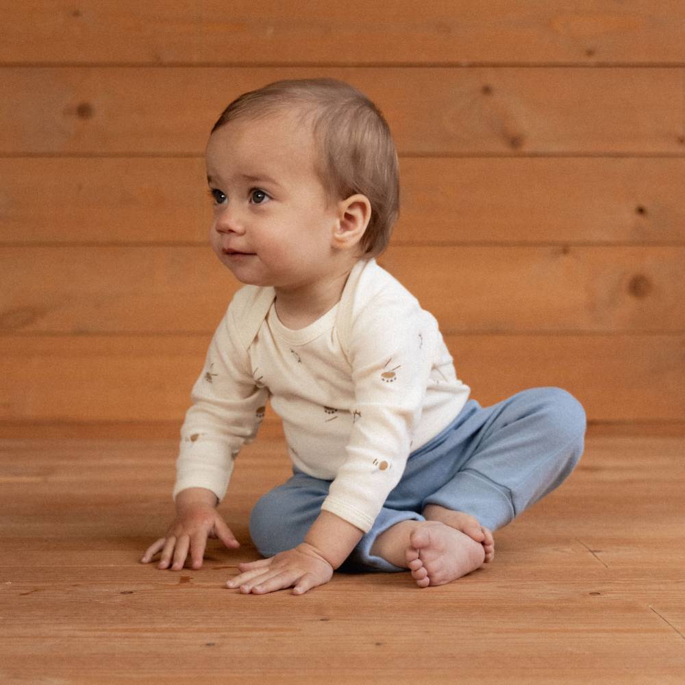 A baby sitting cross-legged on a wooden floor against a wooden plank wall, wearing a cream long-sleeve shirt with small embroidered motifs and light blue pants.