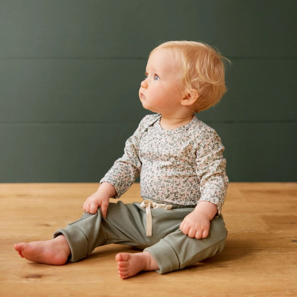 Baby sitting on a wooden floor wearing a floral patterned shirt and green pants with beige drawstring against a dark green wall.