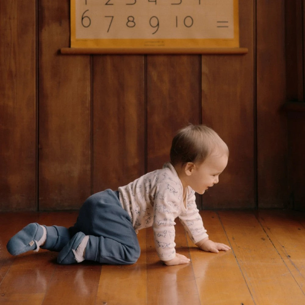 Baby crawling on a wooden floor with navy blue pants and blue booties and a number chart on the wall.
