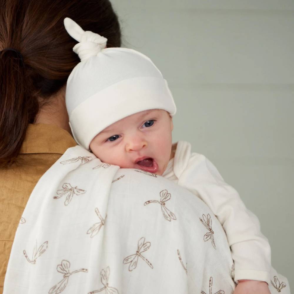 Baby being held over an adults shoulder with dragonfly patterned receiving blanket wearing a white cotton knotted hat 